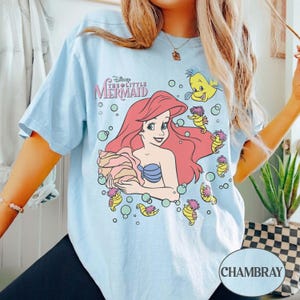 May include: Pale blue t-shirt with a graphic of Ariel from Disney's The Little Mermaid. The design includes Ariel with red hair, a blue seashell bra, and other characters. The text "Disney The Little Mermaid" is above the graphic.