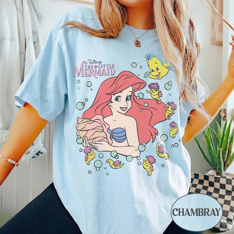 May include: Pale blue t-shirt with a graphic of Ariel from Disney's The Little Mermaid. The design includes Ariel with red hair, a blue seashell bra, and other characters. The text "Disney The Little Mermaid" is above the graphic.