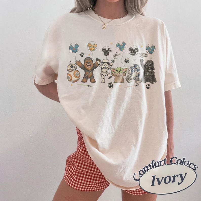 May include: Ivory-colored t-shirt featuring Star Wars characters holding balloon-shaped Mickey Mouse heads. Characters include BB-8, Chewbacca, Stormtrooper, Grogu, R2-D2, and Darth Vader. The shirt has a Comfort Colors label.