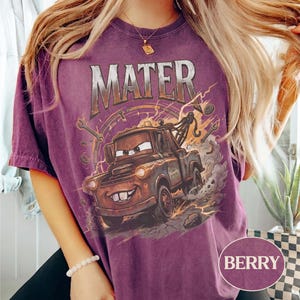 May include: A berry-colored short-sleeve t-shirt featuring a graphic print of the character Mater from the movie Cars. The design includes the text MATER in a metallic font above the brown tow truck, surrounded by wrenches and dust clouds.