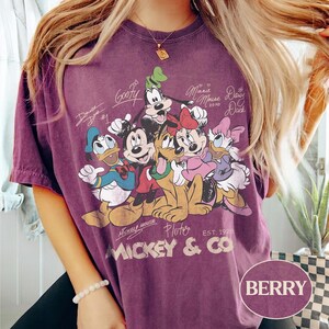 K&ouml;nnte beinhalten: Beerenfarbenes T-Shirt mit einem Aufdruck von Micky Maus und seinen Freunden, darunter Donald Duck, Goofy und Pluto. Das Design enth&auml;lt Unterschriften der Charaktere und den Text "Mickey & Co. Est. 1928". Das Shirt hat einen Vintage-Look.
