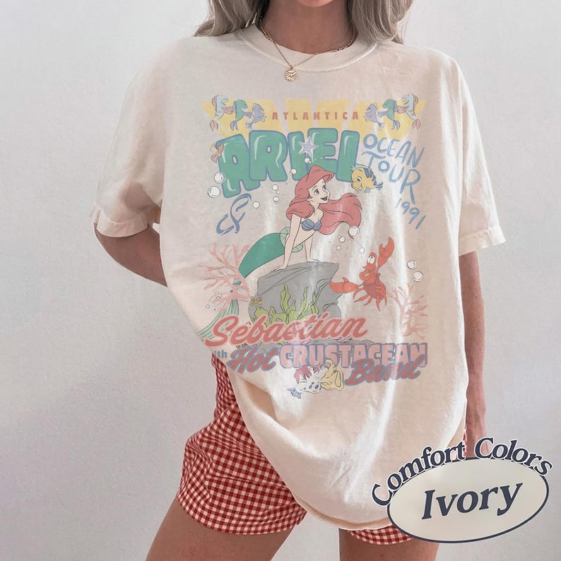 May include: An ivory t-shirt featuring a graphic print of Ariel, Flounder, and Sebastian with text that reads Atlantica Ocean Tour 1991, Sebastian and Hot Crustacean Band. The shirt has a crew neckline and short sleeves, worn with red and white gingham shorts.