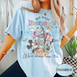 May include: Light blue t-shirt featuring a graphic of Disney characters, including Mickey and Minnie Mouse, Donald and Daisy Duck, Goofy, and Pluto. The shirt has the text "Walt Disney's Disneyworld" and "Where dreams come true."