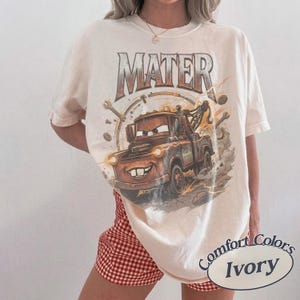 May include: An ivory short-sleeve t-shirt featuring a graphic of the character Mater from the movie Cars. The design includes the text MATER in a metallic font above the brown tow truck, surrounded by wrenches and gears on a dusty background.