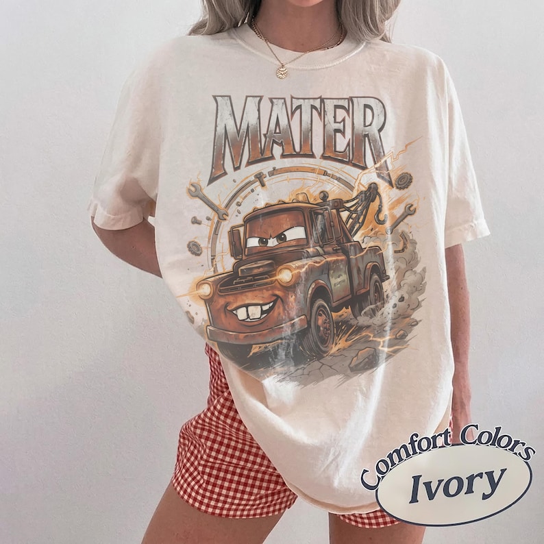 May include: An ivory short-sleeve t-shirt featuring a graphic of the character Mater from the movie Cars. The design includes the text MATER in a metallic font above the brown tow truck, surrounded by wrenches and gears on a dusty background.