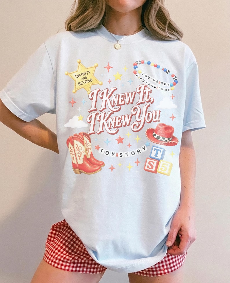May include: A light blue t-shirt featuring a graphic print with the text "I KNEW IT, I KNEW YOU" and "TOY STORY". The design includes a yellow sheriff star, a colorful beaded necklace, red cowboy boots, a red cowboy hat, and alphabet blocks.