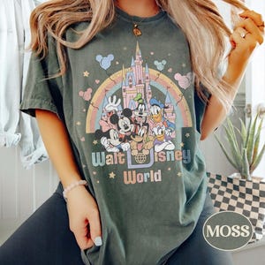 May include: Dark green t-shirt featuring a colorful graphic of Disney characters in front of a castle and rainbow. The text "Walt Disney World" is printed below the graphic. The shirt has a crew neck and short sleeves.
