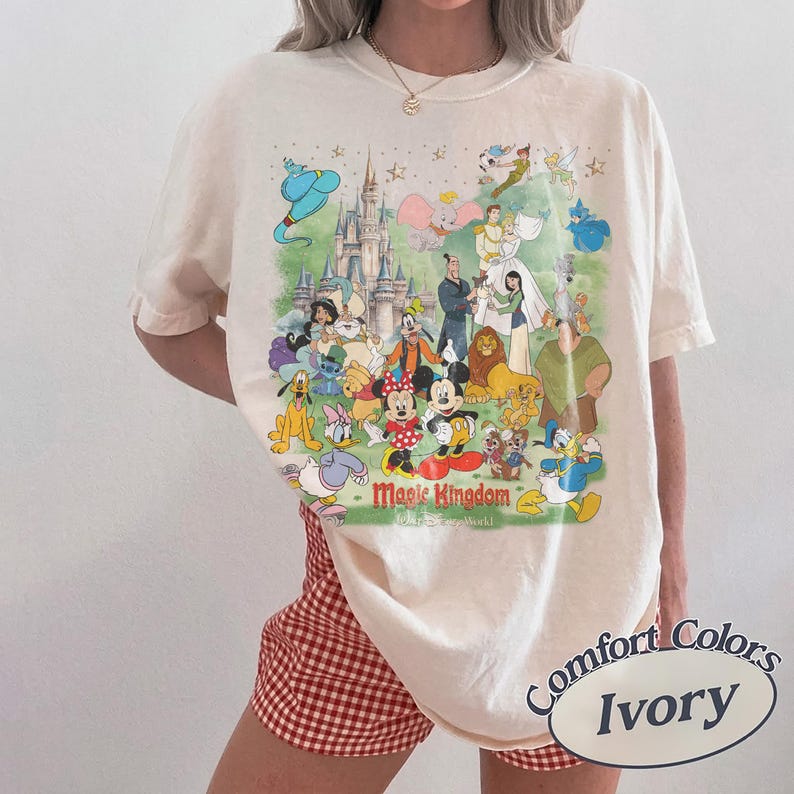May include: Ivory-colored t-shirt featuring a colorful graphic of Disney characters and the text "Magic Kingdom". The design includes Mickey and Minnie Mouse, Aladdin, and other beloved characters. The shirt is labeled "Comfort Colors".