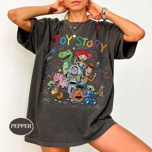 Pode incluir: T-shirt cinza escuro com um gr&aacute;fico colorido dos personagens de Toy Story, incluindo Woody, Buzz Lightyear e Rex. As palavras "Toy Story" est&atilde;o escritas no topo em uma fonte de arco-&iacute;ris. A camisa tem um visual vintage.