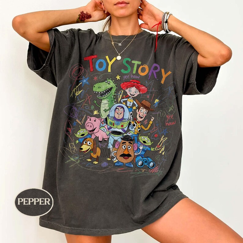 Pode incluir: T-shirt cinza escuro com um gr&aacute;fico colorido dos personagens de Toy Story, incluindo Woody, Buzz Lightyear e Rex. As palavras "Toy Story" est&atilde;o escritas no topo em uma fonte de arco-&iacute;ris. A camisa tem um visual vintage.