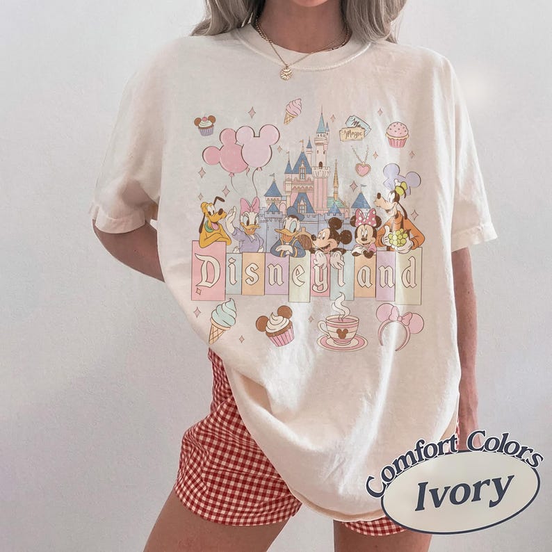 Może przedstawiać: Kremowa koszulka z grafiką Disneyland z postaciami z kresk&oacute;wek i zamkiem. Projekt zawiera słowo "Disneyland" w pastelowej czcionce, a także babeczki, lody i balony. Koszulka jest oznaczona jako "Comfort Colors Ivory".