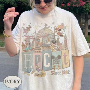 May include: An ivory short-sleeve t-shirt featuring a graphic print of Epcot with Disney characters including Mickey Mouse, Minnie Mouse, Donald Duck, and Pluto. The design includes the text EPCOT and Since 1982 with various park landmarks.