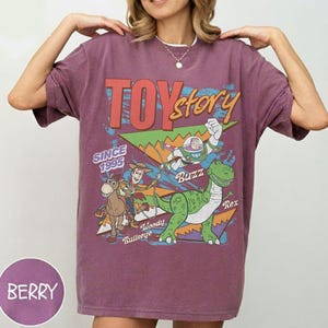May include: Berry-colored t-shirt featuring a graphic from the movie "Toy Story." The design includes the text "Toy Story" and the characters Buzz Lightyear, Woody, Rex, and Bullseye. The shirt also has the text "Since 1995."