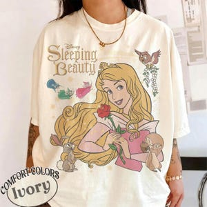 May include: An ivory t-shirt featuring a graphic print of Sleeping Beauty with long blonde hair, holding a red rose. The design includes small forest animals, fairies, and the text Disney Sleeping Beauty. The shirt has a relaxed, loose fit.