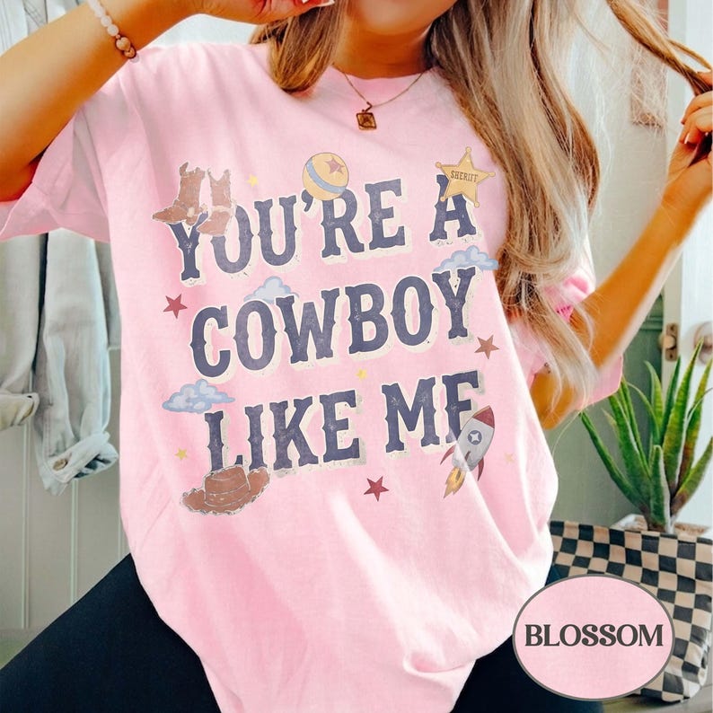 May include: A light pink short-sleeve t-shirt featuring the dark blue text "YOU'RE A COWBOY LIKE ME" surrounded by graphic illustrations of cowboy boots, a sheriff star, a ball, clouds, stars, and a rocket ship in various colors.