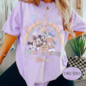 May include: Orchid-colored t-shirt featuring a graphic of Disney characters, a rainbow, and a castle. The text "Walt Disney World" is printed below the characters. The shirt has short sleeves.