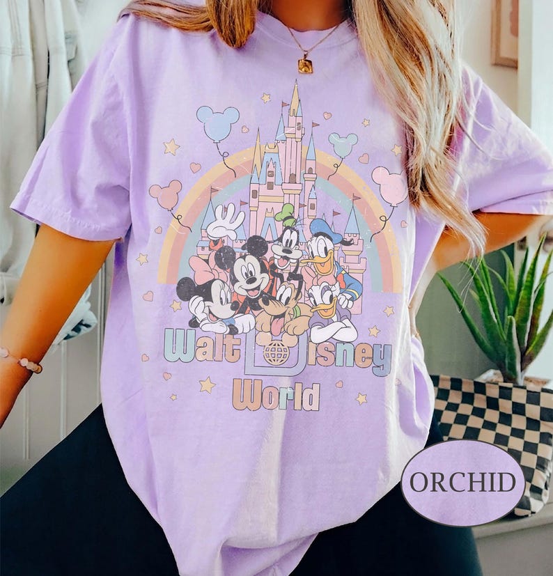 May include: Orchid-colored t-shirt featuring a graphic of Disney characters, a rainbow, and a castle. The text "Walt Disney World" is printed below the characters. The shirt has short sleeves.
