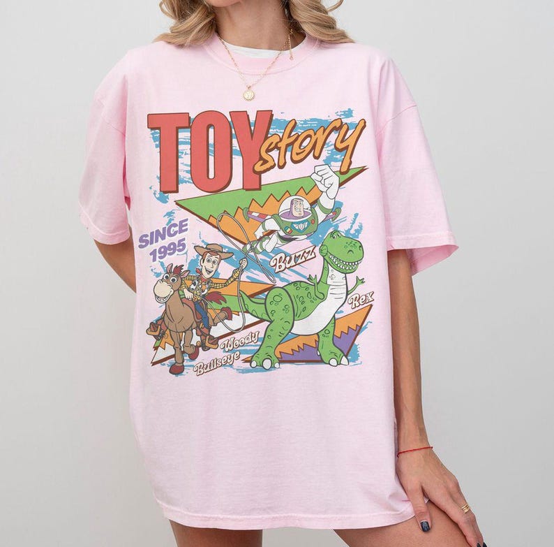 May include: A light pink t-shirt featuring a colorful graphic from the movie Toy Story. The design includes the text "TOY STORY" and images of Woody, Buzz Lightyear, Rex, and Bullseye. The text "SINCE 1995" is also included.