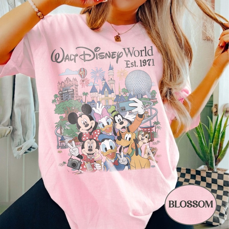 May include: A light pink t-shirt featuring a graphic print of Walt Disney World Est. 1971. The design includes illustrations of Mickey Mouse, Minnie Mouse, Donald Duck, Daisy Duck, Goofy, Pluto, and Chip 'n Dale in front of park landmarks.