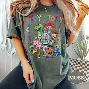 Pode incluir: Camiseta verde musgo com as palavras "TOY STORY" em letras coloridas sobre um gr&aacute;fico de personagens do filme de anima&ccedil;&atilde;o. O design inclui Woody, Buzz Lightyear, Rex e outros personagens. A camisa &eacute; de estilo casual.