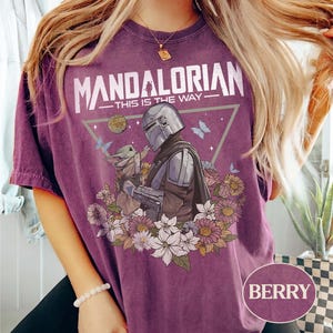 Op de afbeelding: Bessenkleurig T-shirt met een afbeelding van The Mandalorian en Grogu, omringd door bloemen en vlinders. De tekst "MANDALORIAN - THIS IS THE WAY" staat boven de afbeelding. Het shirt heeft een relaxte pasvorm.