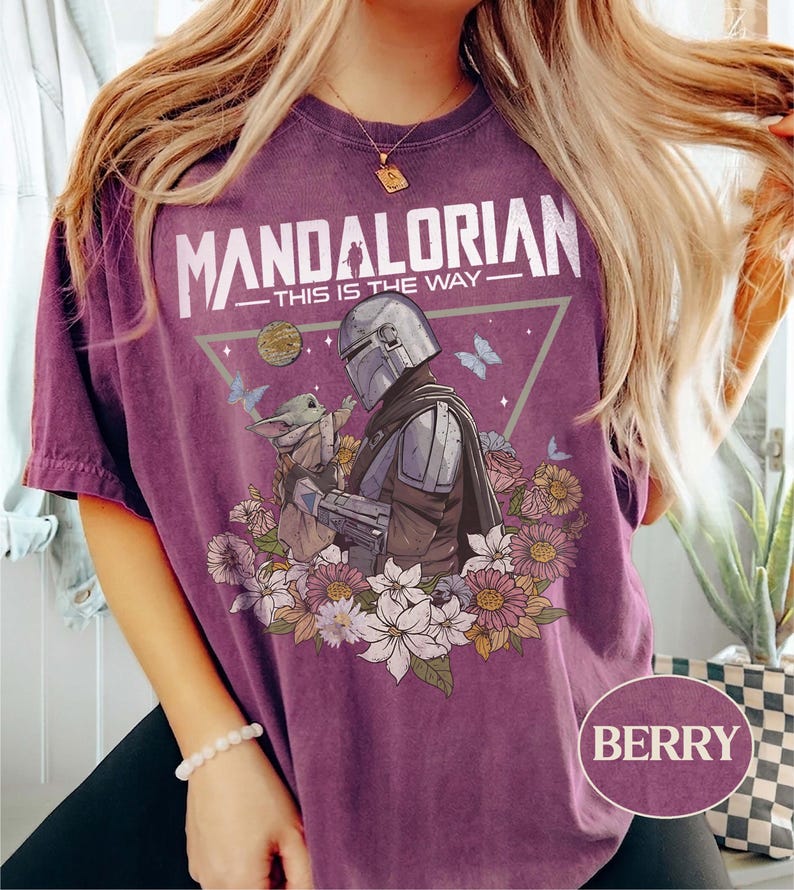 Op de afbeelding: Bessenkleurig T-shirt met een afbeelding van The Mandalorian en Grogu, omringd door bloemen en vlinders. De tekst "MANDALORIAN - THIS IS THE WAY" staat boven de afbeelding. Het shirt heeft een relaxte pasvorm.