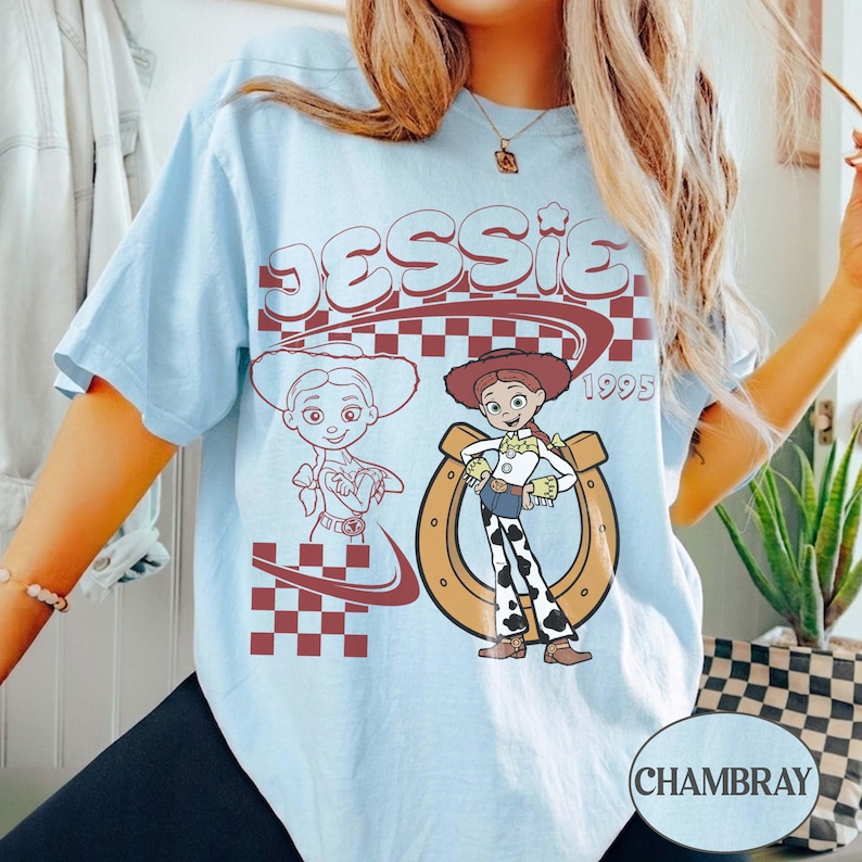 May include: A light blue t-shirt featuring a graphic print of the character Jessie. The design includes the text JESSIE and 1995, a brown horseshoe, a red and white checkered pattern, and a line-art illustration of the character on the left.