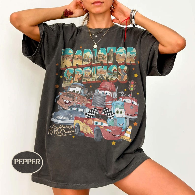 K&ouml;nnte beinhalten: Dunkelgraues T-Shirt mit einem farbenfrohen Aufdruck von Charakteren aus dem Film Cars, mit dem Text "Radiator Springs" und "Lightning McQueen". Das Design zeigt verschiedene Autocharaktere und Sterne.