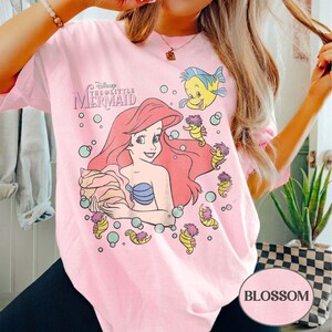 May include: Pale pink t-shirt with a graphic of Ariel from Disney's The Little Mermaid. The design includes Ariel, Flounder, seahorses, and bubbles. The text "Disney The Little Mermaid" is above Ariel. The word "Blossom" is in a white oval.