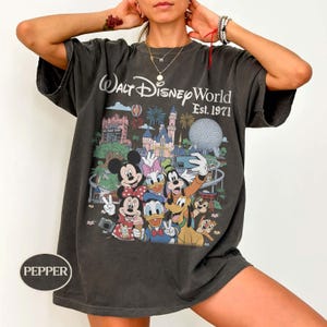 May include: A charcoal gray short-sleeve t-shirt featuring a graphic print of Walt Disney World Est. 1971 with various cartoon characters including Mickey Mouse, Minnie Mouse, Donald Duck, Daisy Duck, Goofy, Pluto, and Chip 'n Dale in front of the park.