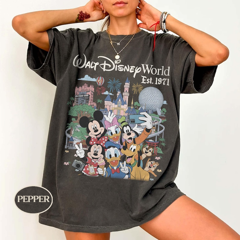 May include: A charcoal gray short-sleeve t-shirt featuring a graphic print of Walt Disney World Est. 1971 with various cartoon characters including Mickey Mouse, Minnie Mouse, Donald Duck, Daisy Duck, Goofy, Pluto, and Chip 'n Dale in front of the park.