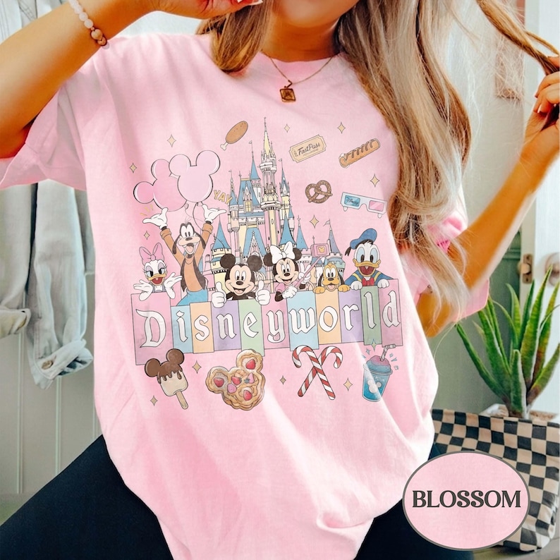 May include: Pale pink t-shirt with a Disney World graphic. The design features Mickey and Minnie Mouse, Donald Duck, and other characters in front of a castle. The word "Disneyworld" is written across the bottom in colourful letters.
