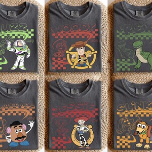 May include: Six charcoal gray t-shirts featuring Toy Story characters Buzz Lightyear, Woody, Rex, Mr. Potato Head, Jessie, and Slinky Dog. Each shirt displays the character name, a 1995 date, a checkered pattern, and a line art illustration.