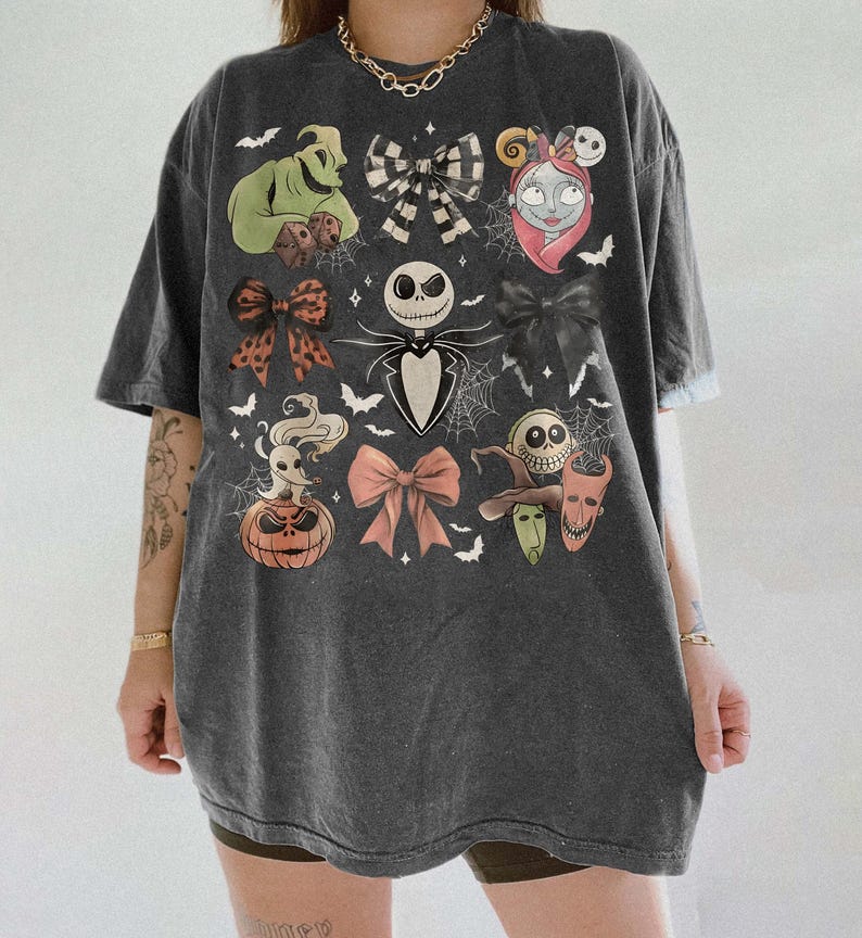 May include: A charcoal gray oversized t-shirt featuring a graphic print of characters from The Nightmare Before Christmas, including Jack Skellington, Sally, Zero, and Oogie Boogie, interspersed with various bows, spiderwebs, and bats.