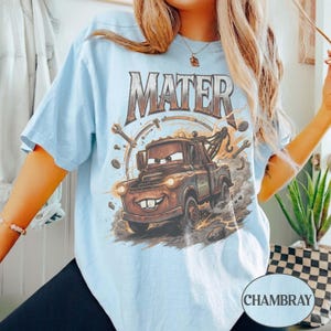 May include: A light blue t-shirt featuring a graphic print of a brown tow truck character with the text MATER above it. The design includes illustrations of mechanical tools like wrenches and gears surrounding the truck on a dusty background.