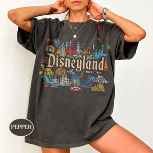Op de afbeelding: Donkergrijs t-shirt met een kleurrijke Disneyland Resort afbeelding. Het ontwerp toont iconische bezienswaardigheden en het Disneyland Resort logo. Het shirt heeft korte mouwen en een relaxte pasvorm.