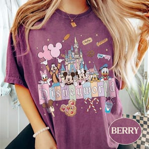May include: Berry-coloured t-shirt with a Disney World graphic. The design features Mickey and Minnie Mouse, Donald Duck, Goofy, and other characters, a castle, and the word "Disneyworld". The shirt has a vintage, washed appearance.