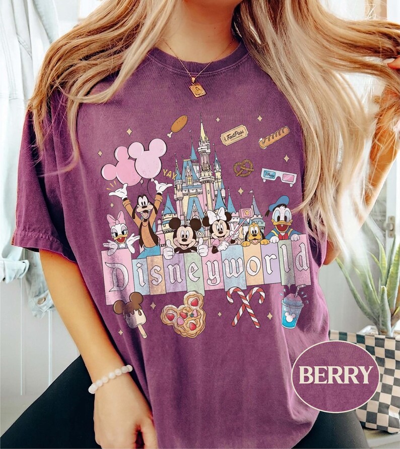 May include: Berry-coloured t-shirt with a Disney World graphic. The design features Mickey and Minnie Mouse, Donald Duck, Goofy, and other characters, a castle, and the word "Disneyworld". The shirt has a vintage, washed appearance.