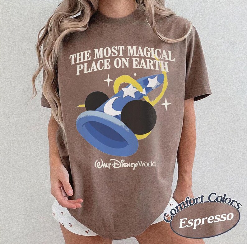 Puede incluir: Camiseta color espresso con el texto "THE MOST MAGICAL PLACE ON EARTH" y un gr&aacute;fico de un sombrero de mago azul con estrellas. El logotipo de Walt Disney World est&aacute; debajo. De la colecci&oacute;n Comfort Colors.