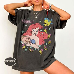 May include: Dark grey t-shirt with a graphic of Ariel from Disney's The Little Mermaid. Ariel has red hair and a blue seashell bra. The shirt also features Flounder and seahorse illustrations. The text "The Little Mermaid" is above Ariel.
