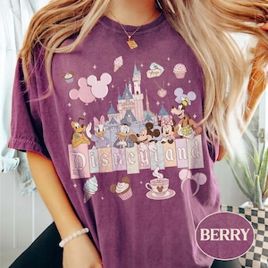 Może przedstawiać: Koszulka w kolorze jagodowym z grafiką Disneyland. Projekt zawiera zamek Disneyland, Myszka Miki i Minnie, Kaczor Donald, Daisy Duck, Goofy i Pluto. Słowo "Disneyland" jest wyświetlane na dole.