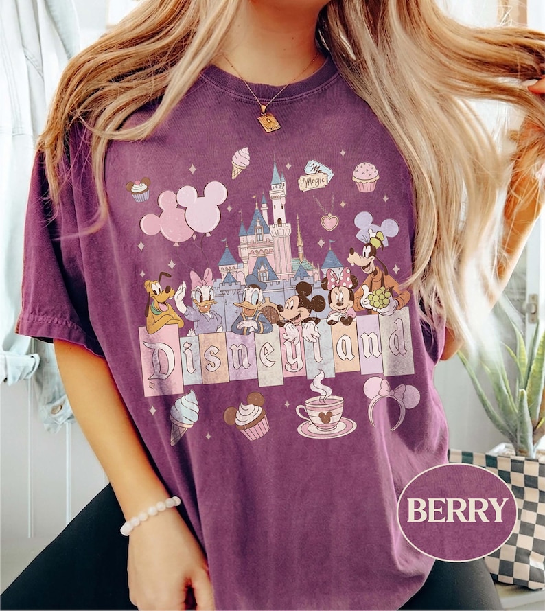 Może przedstawiać: Koszulka w kolorze jagodowym z grafiką Disneyland. Projekt zawiera zamek Disneyland, Myszka Miki i Minnie, Kaczor Donald, Daisy Duck, Goofy i Pluto. Słowo "Disneyland" jest wyświetlane na dole.
