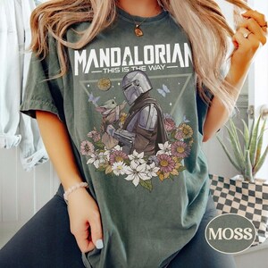Op de afbeelding: Mosgroen t-shirt met een afbeelding van The Mandalorian en Grogu, omringd door bloemen en vlinders. De tekst "MANDALORIAN - THIS IS THE WAY" staat bovenaan gedrukt. Een rond "MOSS" label staat rechtsonder.