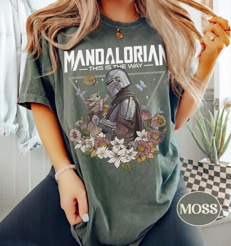Op de afbeelding: Mosgroen t-shirt met een afbeelding van The Mandalorian en Grogu, omringd door bloemen en vlinders. De tekst "MANDALORIAN - THIS IS THE WAY" staat bovenaan gedrukt. Een rond "MOSS" label staat rechtsonder.