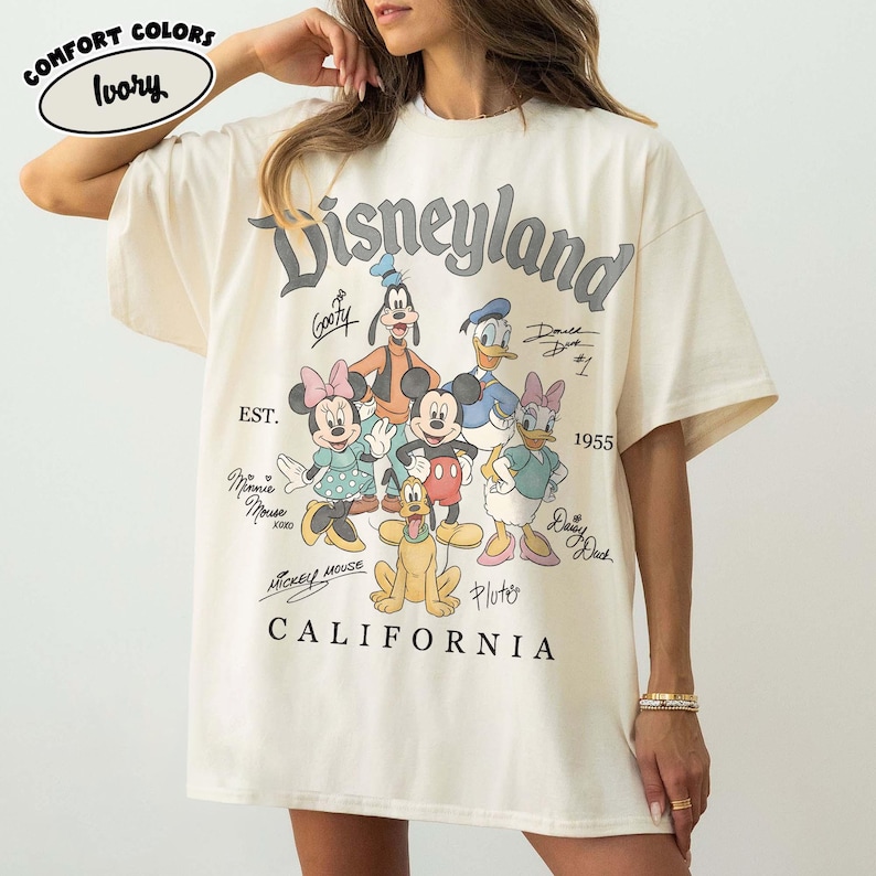 May include: Ivory-coloured t-shirt with the word "Disneyland" above a graphic of Disney characters including Mickey Mouse, Minnie Mouse, Goofy, Donald Duck, and Pluto. The text "CALIFORNIA" is printed below the graphic. The shirt is labelled "COMFORT COLORS".