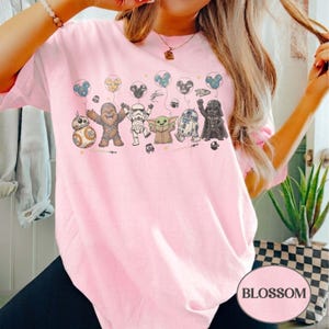 May include: A light pink t-shirt featuring a graphic of Star Wars characters holding balloons. The characters include BB-8, Chewbacca, a Stormtrooper, Grogu, R2-D2, and Darth Vader. The balloons have Mickey Mouse ears. The word "BLOSSOM" is in a white oval.