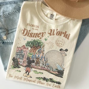 May include: Cream-coloured t-shirt with a graphic of Disney World in Florida. The design includes Mickey Mouse, the Hollywood Tower, Cinderella's castle, and Spaceship Earth. Text on the shirt reads "Disney World" and "The Most Magical Place On Earth."