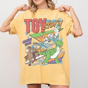 May include: Yellow t-shirt featuring a graphic from the movie Toy Story. The design includes the text "TOY STORY" in red, along with images of Woody, Buzz Lightyear, and Rex. The text "SINCE 1995" is also included.