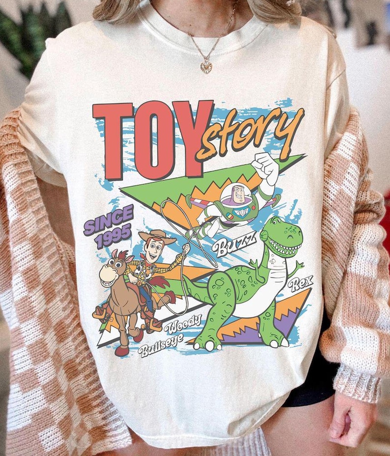May include: Cream-colored t-shirt featuring a graphic design from the movie "Toy Story." The design includes the text "TOY Story" in red and yellow, along with images of Woody, Buzz Lightyear, Rex, and Bullseye. The text "SINCE 1995" is also present.