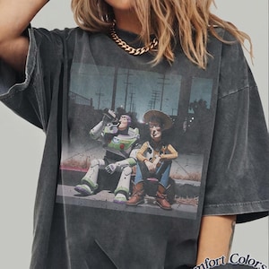 May include: A charcoal grey short-sleeve t-shirt featuring a graphic print of Buzz Lightyear and Woody sitting on a curb. The shirt has a washed-out, vintage appearance. The text "Comfort Colors Pepper" is printed in the bottom right corner.